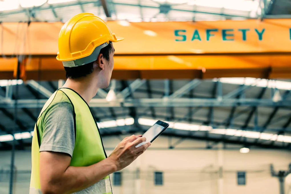 Construction worker wearing yellow hard hat and safety vest using mobile device under safety signage